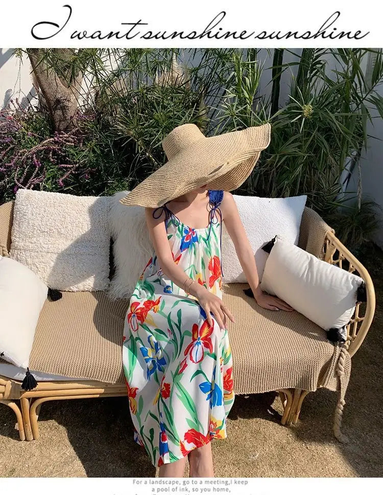 A woman wears a vibrant floral mesh maxi dress with a retro slip silhouette featuring thin straps and a flowing skirt paired with a wide-brimmed straw hat.