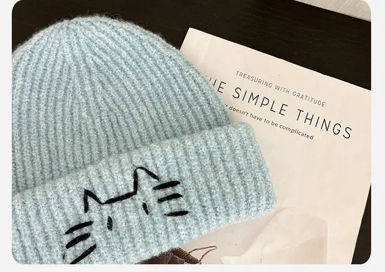 Light blue knitted beanie hat with a black embroidered cat face design made from soft yarn.