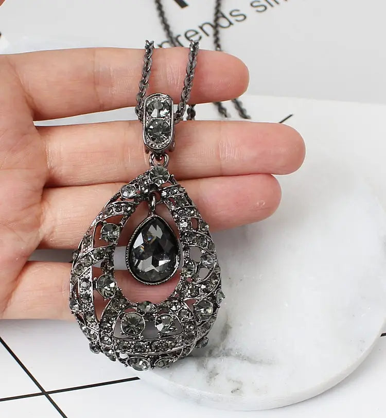 An ornate black teardrop pendant necklace featuring blue filigree metalwork and sparkling dark gemstones on a Korean style sweater chain.