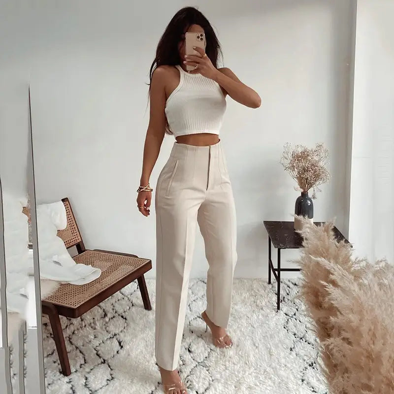 Une femme portant un pantalon taille haute à plis plats et jambes droites de couleur crème, assorti à un top court blanc côtelé.