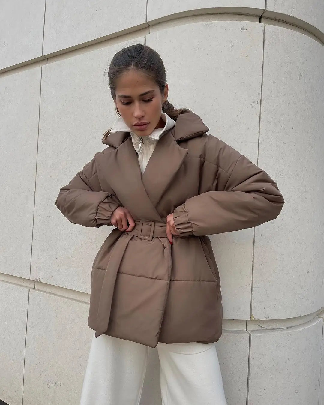 Eine taupefarbene Damen-Gurtsteppjacke mit Kapuze, wattierte Pufferjacke mit Taillengürtel, für den Gebrauch als Winterjacke.