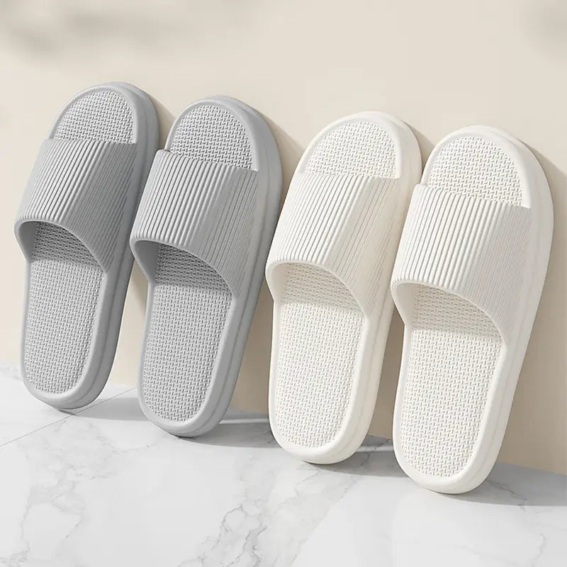 A pair of unisex ribbed anti slip Eva house slippers in soft gray and white with a lightweight minimalist design and a textured rubber sole for grip.