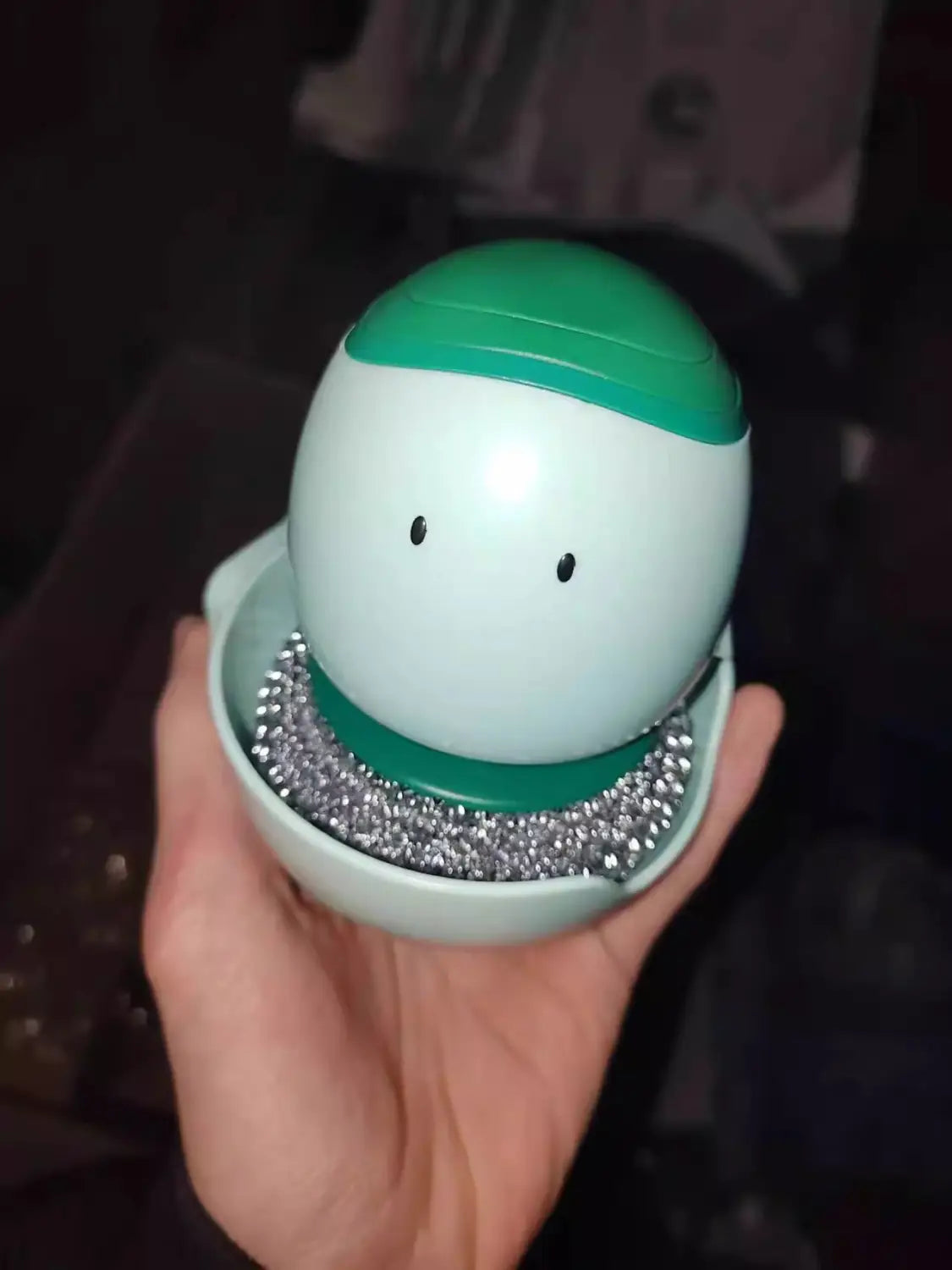 brosse pour plat en forme d’œuf récurant-distributeur automatique de savon liquide en vert clair et blanc, avec un design de visage souriant et un capuchon amovible qui révèle une fine laine d’acier argentée à l’intérieur.