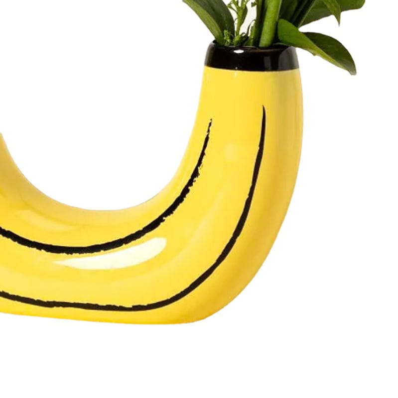 A glossy yellow ceramic banana-shaped vase with black curved lines and a black rim holding green foliage featured as banana ceramic vase yellow fruit shaped home decor.