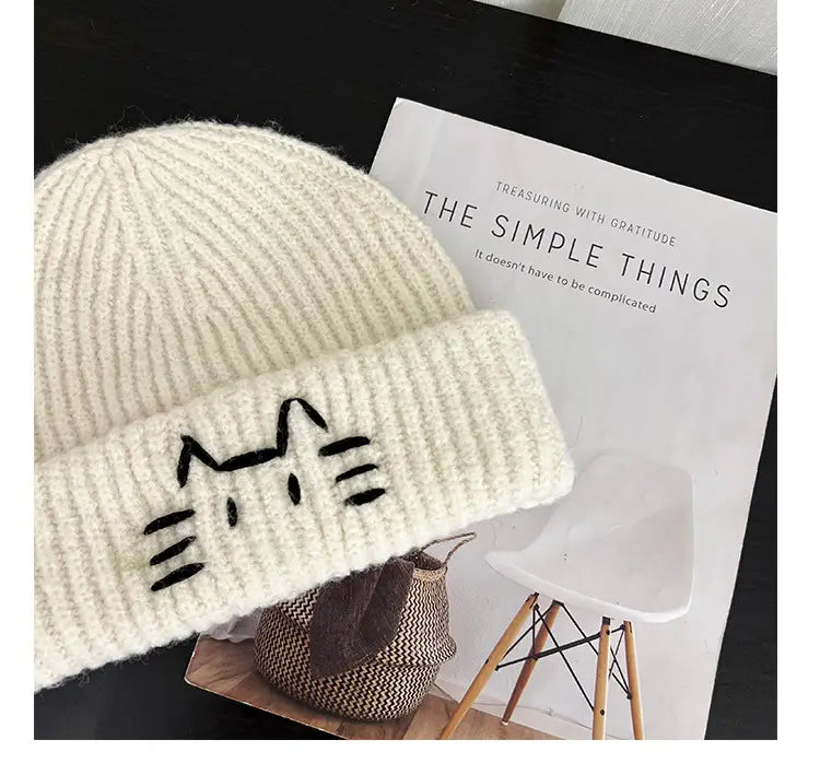 Cream-colored knitted beanie hat featuring a black embroidered cat face design on the folded brim.
