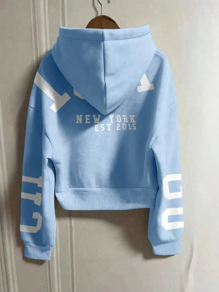 Sweat-shirt à capuche zippé court bleu clair, avec le texte « New York Est 2015 » blanc et de grandes lettres sur les manches.