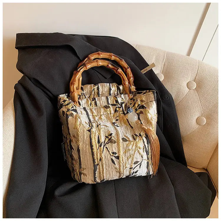 A stylish bamboo handbag with tassel featuring beige and gold textured fabric and an elegant tree pattern forest canvas.