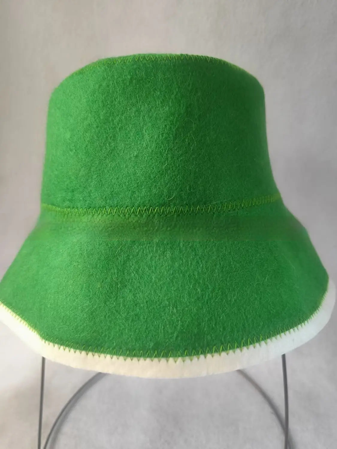 Eine kräftige grüne Wollfilz-Fischerhut-Kappe mit weißem Zierbesatz und sichtbaren Nähten entlang des Randes, als faltbares Winteraccessoire konzipiert.
