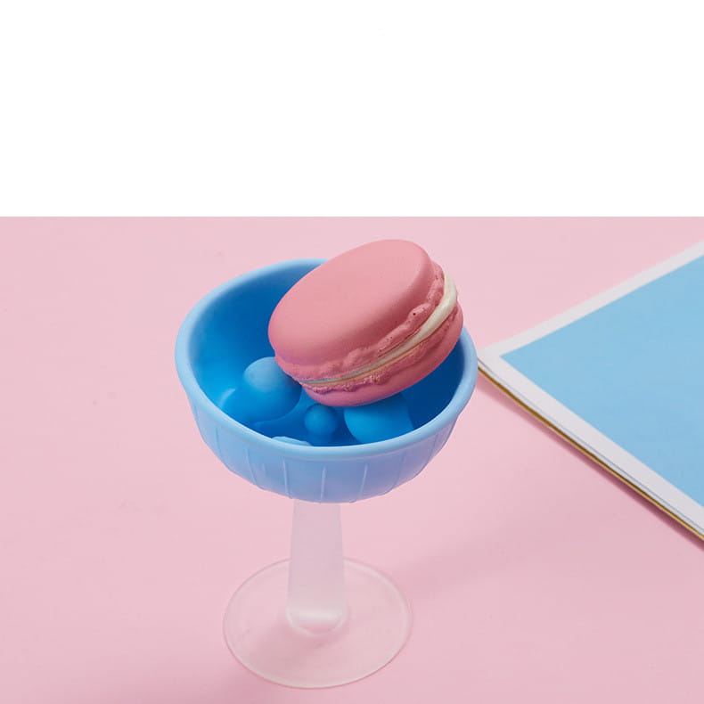 a pink macaron with white filling displayed in a translucent blue plastic cocktail glass featuring a silicone baby teether and pig shape toothbrush holder