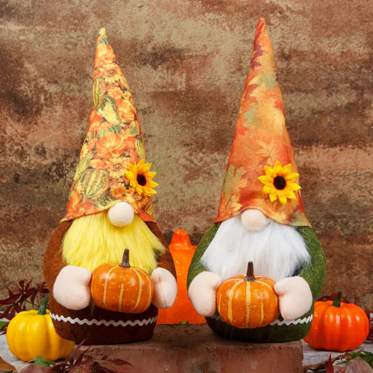 Zwei kuschelige Herbst-Danksagungs-Gnom-Plüschfiguren: eine mit braunem Körper und gelbem Bart, die einen orangefarbenen Kürbis hält, die andere mit grünem Körper und weißem Bart, die ebenfalls einen orangefarbenen Kürbis in den Armen trägt; beide tragen hohe, spitze Hüte, die mit Mustern aus herbstlicher Vegetation und Sonnen verziert sind.