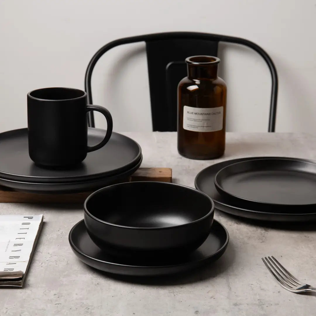 Un juego de vajilla de gres mate negro de 12 piezas para tres, que presenta platos, tazones y una taza de cerámica mate negra con diseño minimalista.
