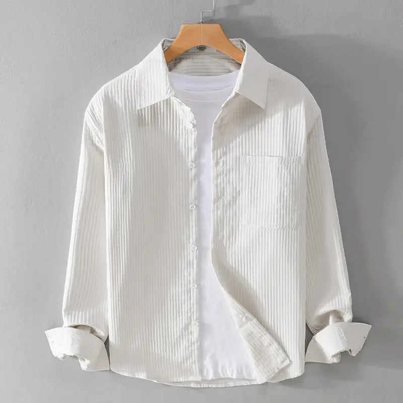 Chemise homme décontractée à manches longues blanche, présentant une texture côtelée verticale et une poche poitrine, suspendue à un cintre en bois.