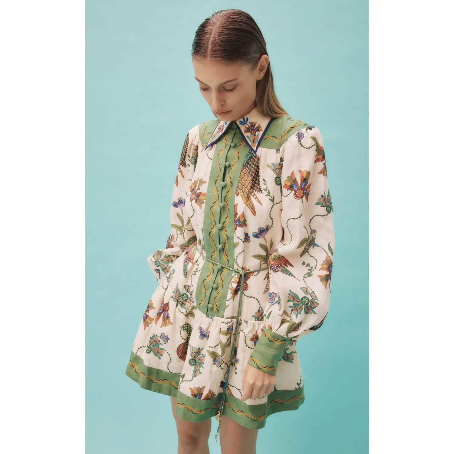 A cream-colored botanical print puff sleeve midi dress with long sleeves, intricate bird and botanical design in earthy tones, and a green corded front panel and matching cuffs.