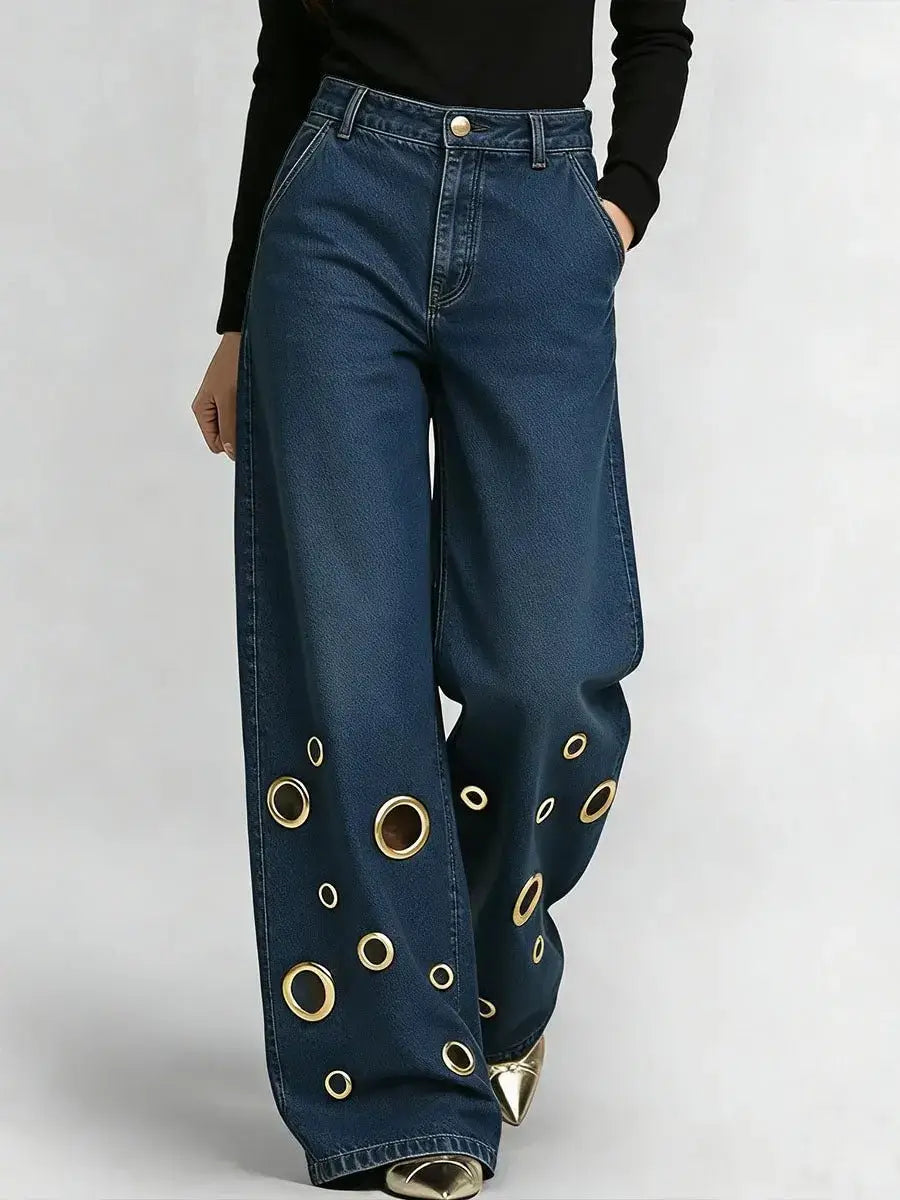 Jeans en denim bleu foncé à œillets et jambes larges, avec des œillets circulaires décoratifs ton sur ton or le long des bas de jambes.