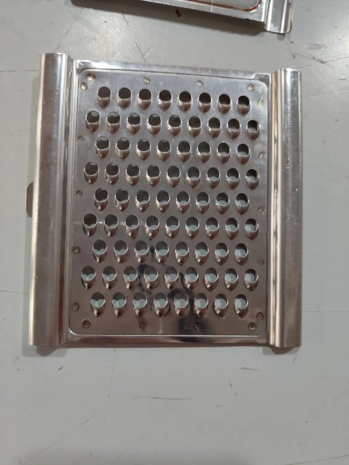 Juego de rallador y cortador multiusos de acero inoxidable con un rallador rectangular con orificios redondeados espaciados uniformemente y bordes laterales elevados.