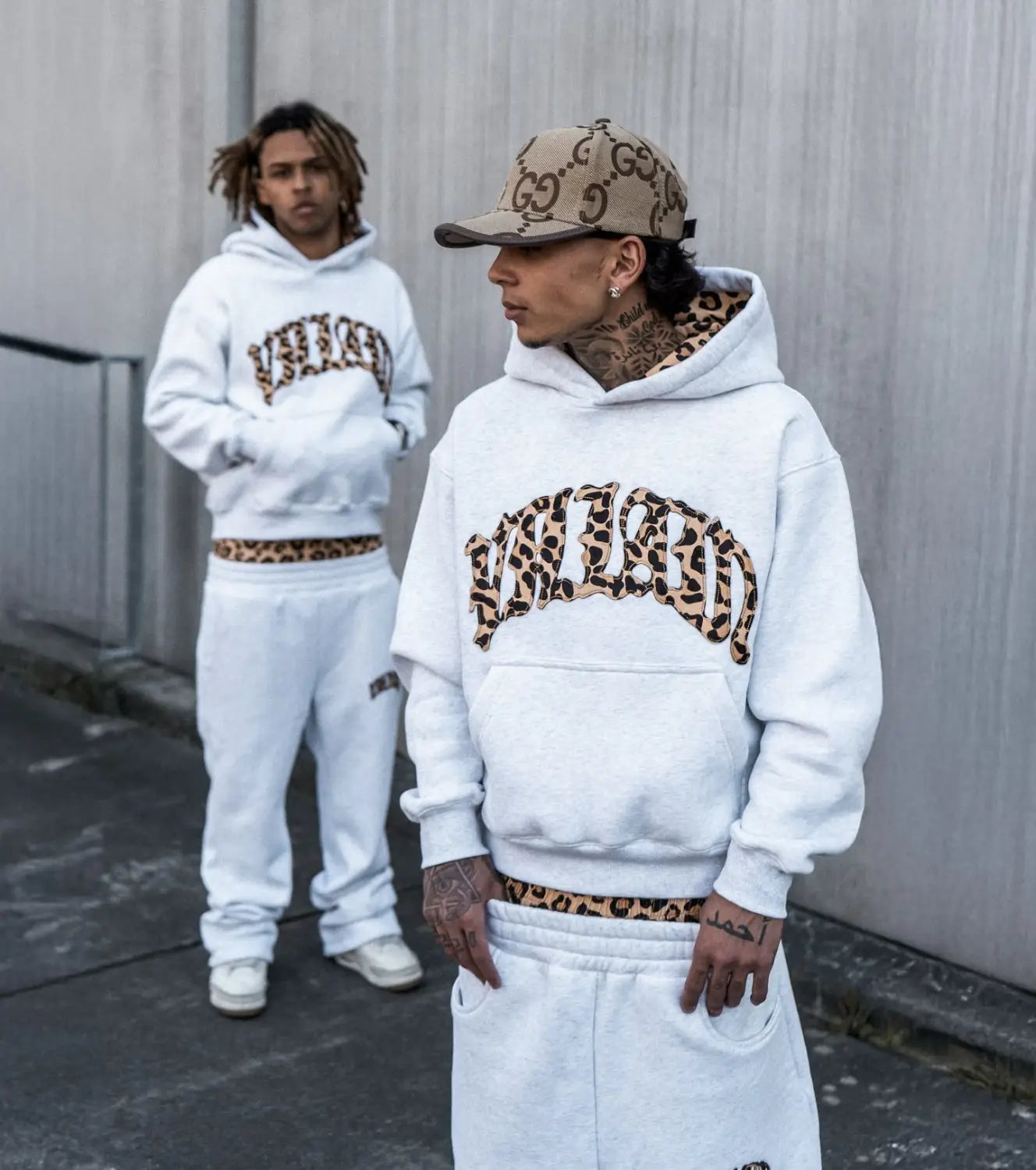 Une personne portant un sweat à capuche blanc avec des inscriptions à imprimé léopard sur la poitrine et une ceinture assortie, issue de l’ensemble sweat à capuche et jogging à imprimé léopard, se tient devant une autre personne habillée de manière similaire.
