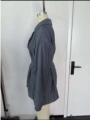 Grauer Trenchcoat mit Taillengürtel und Pattentaschen, an einer Schaufensterpuppe für Damen-Gurtsteppjacke mit Kapuze, wattierte Pufferjacke für den Winter dargestellt.
