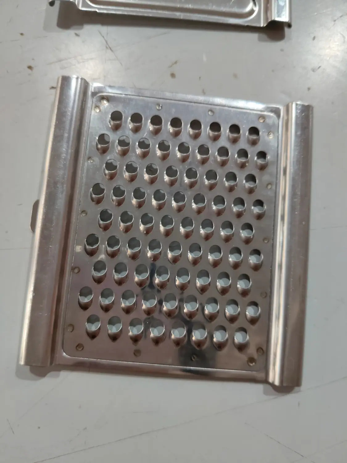 Juego de rallador y cortador multiusos de acero inoxidable con un rallador rectangular de acero inoxidable con orificios circulares espaciados uniformemente y asas laterales elevadas.
