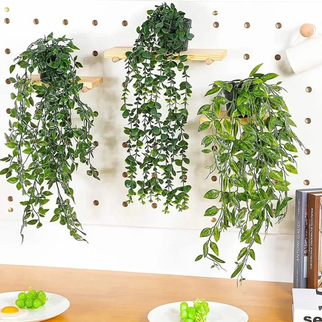 Three artificial hanging plants 3 pack of fake trailing greenery with glossy green leaves hang from wooden shelves mounted on a white pegboard wall.