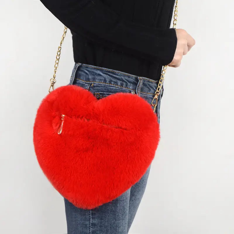 A vibrant red heart shaped crossbody bag made of plush faux fur with a gold tone zipper and chain strap.