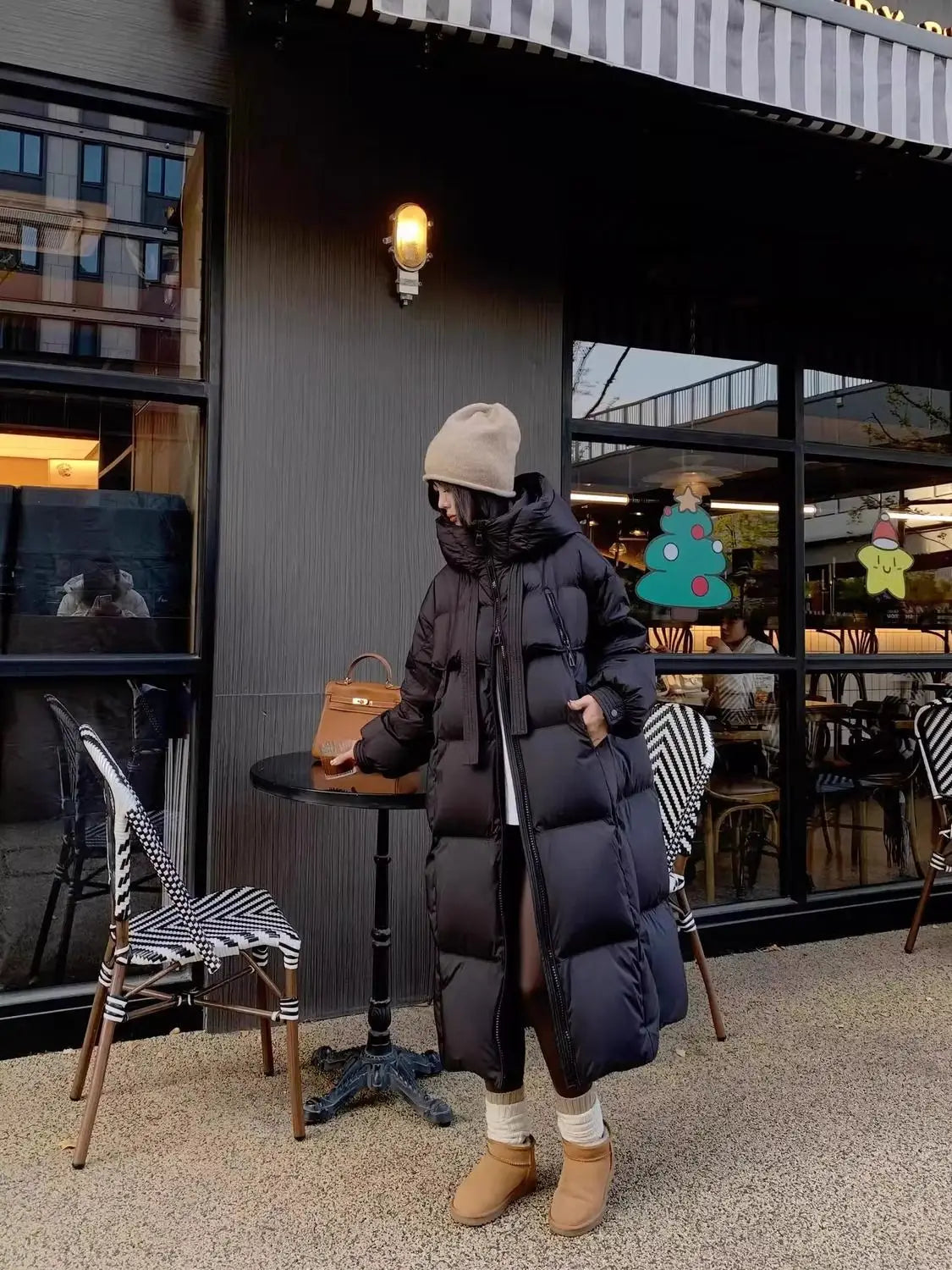 A tan leather handbag with a top handle and gold tone hardware placed on a black table alongside a women long padded down jacket quilted coat.