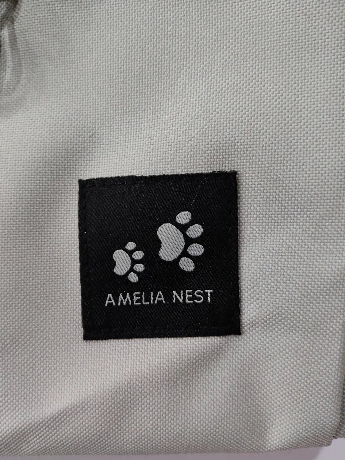 black rectangular label with white paw prints and amelia nest text stitched onto light gray textured fabric of breathable dog backpack mesh pet carrier gray