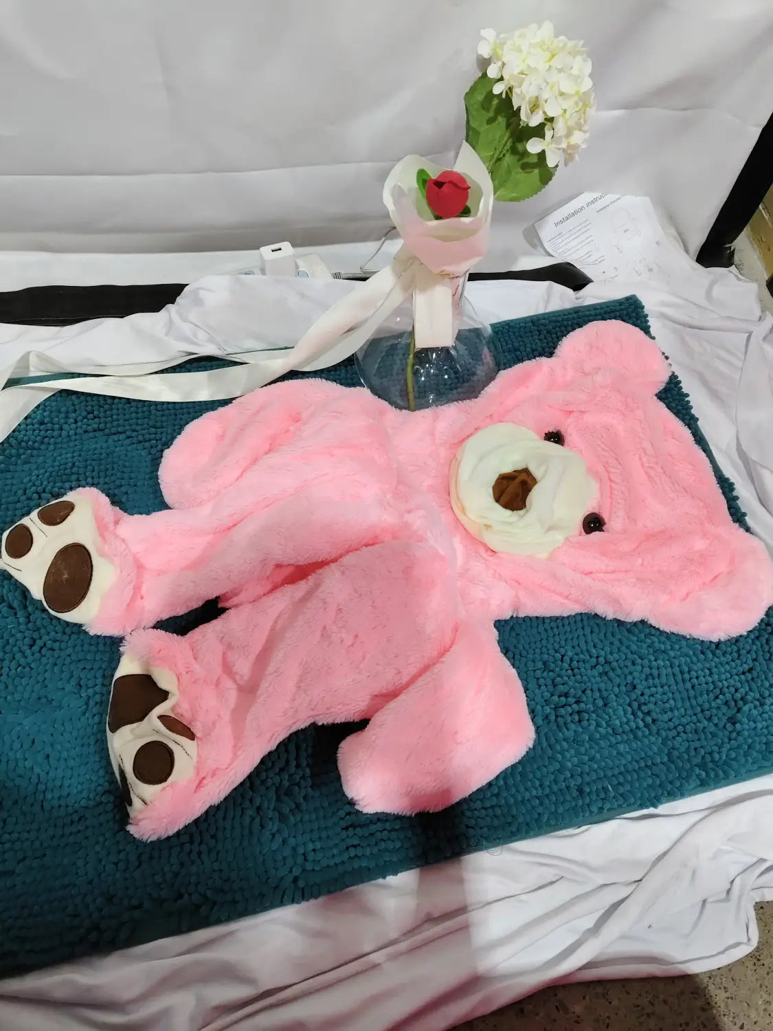 A plush pink giant teddy bear plush toy with white paw pads and a cream-colored snout lies unfilled on a teal mat with a leather shell.