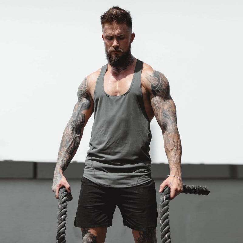 A man wearing a dark gray men’s go hard racerback athletic tank top and black shorts with extensive tattoos on his arms.