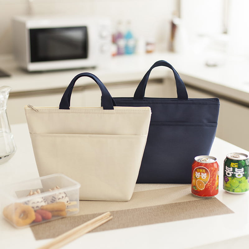 Hellcremefarbene isolierte Oxford-Bento-Lunchbag mit dunkelblauen Griffen und einer vorderen Tasche zum Transport von Mahlzeiten, mit Reißverschlussverschluss.