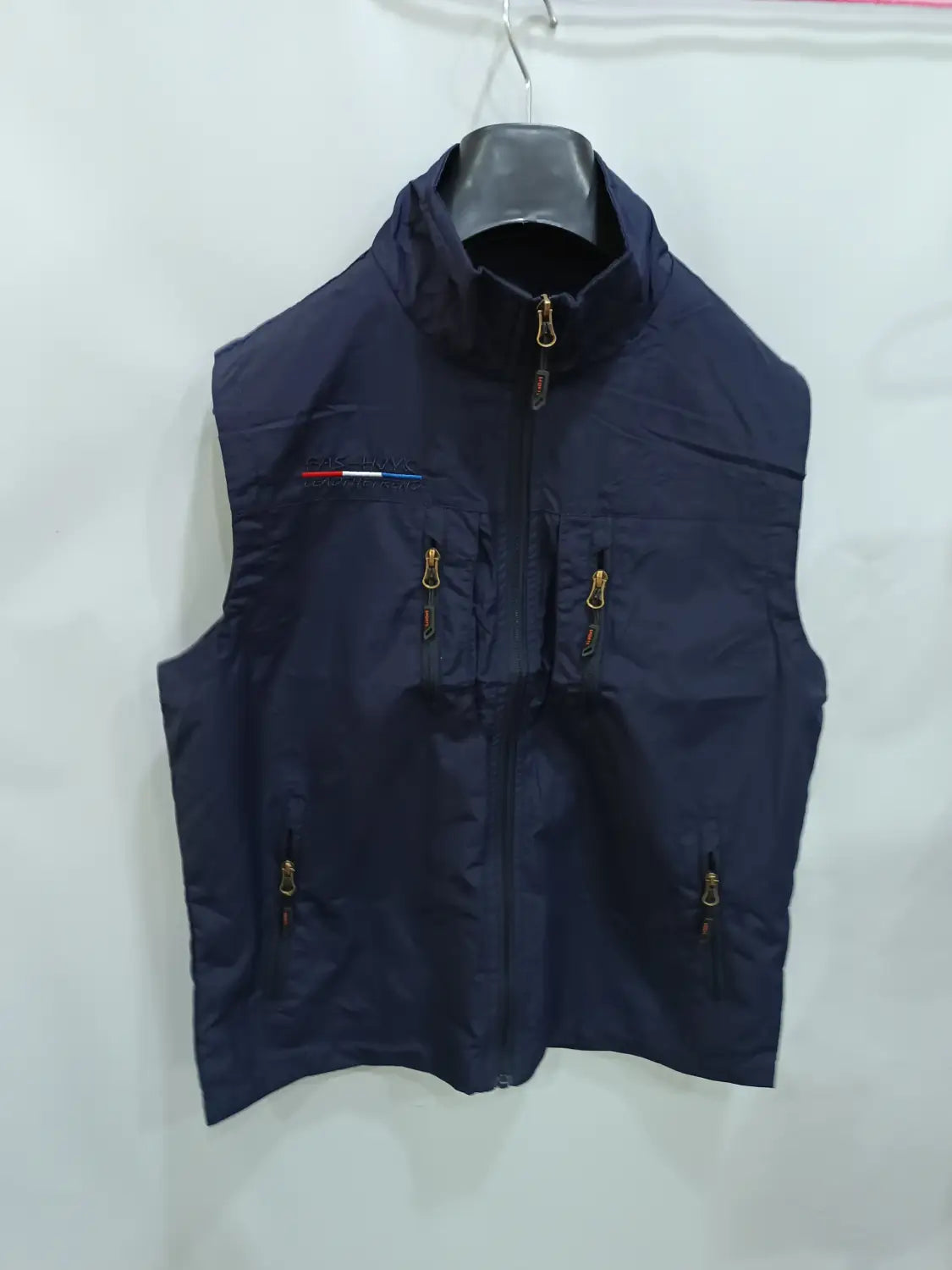 Chaleco utilitario sin mangas con capucha y múltiples bolsillos para hombre en azul marino oscuro, con cremalleras con acabado dorado y un pequeño detalle de franja roja, blanca y azul en el lado izquierdo del pecho.