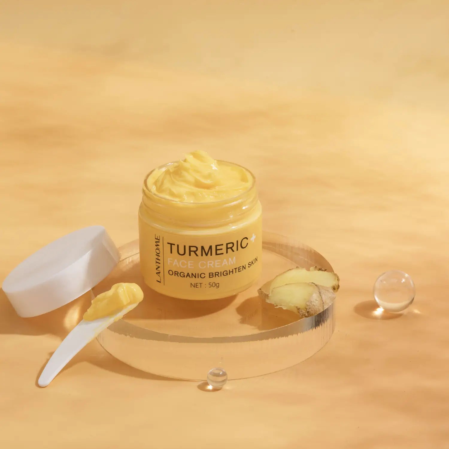 Crème pour le visage au curcuma jaune dans un pot en verre transparent, avec couvercle blanc, portant l’étiquette crème pour le visage au curcuma lanthomé, biologique, illuminer la peau, aux côtés d’une petite cuillère et de gingembre frais, dans l’ensemble de soins au curcuma : huile éclaircissante et crème pour le visage.