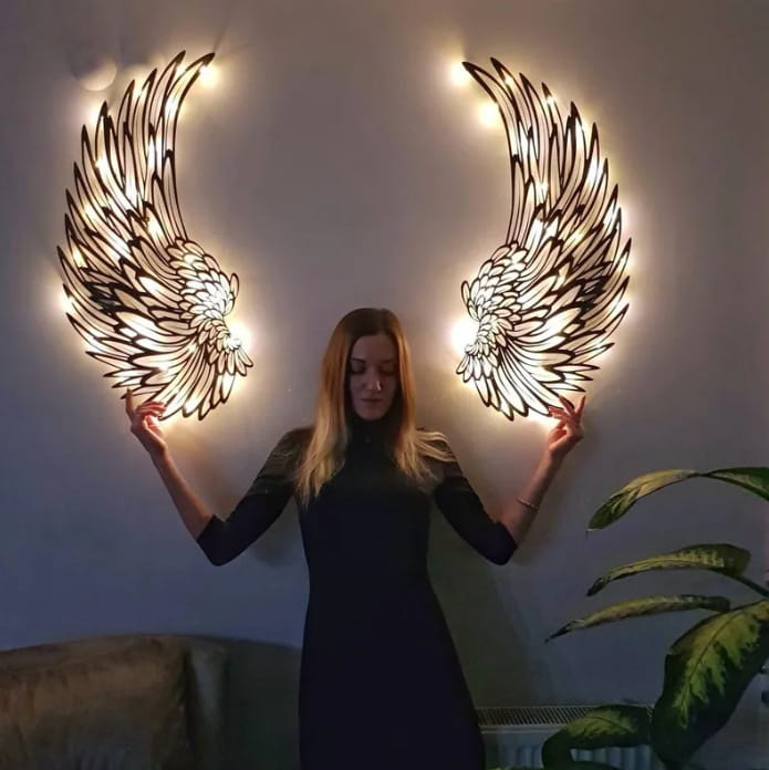 A pair of illuminated metal angel wings wall art with intricate feather details and warm LED lights along the edges.
