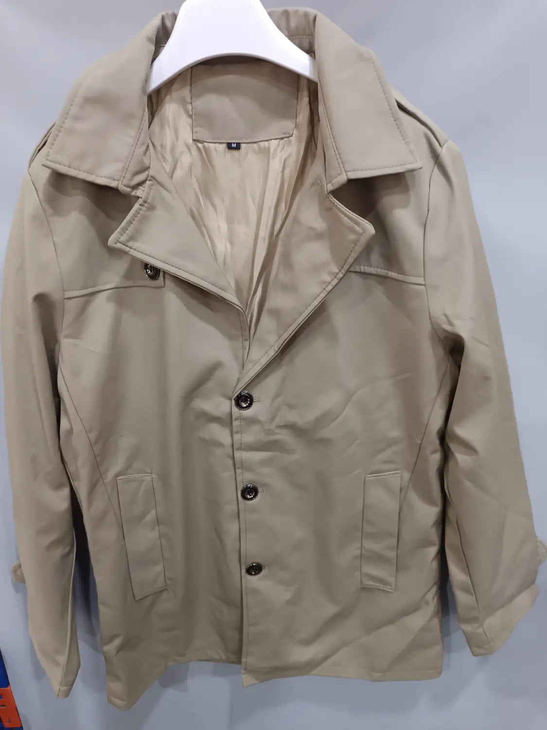 Trench-coat d’affaires homme slim beige avec des boutons noirs et des poches à rabat, suspendu sur un cintre blanc.