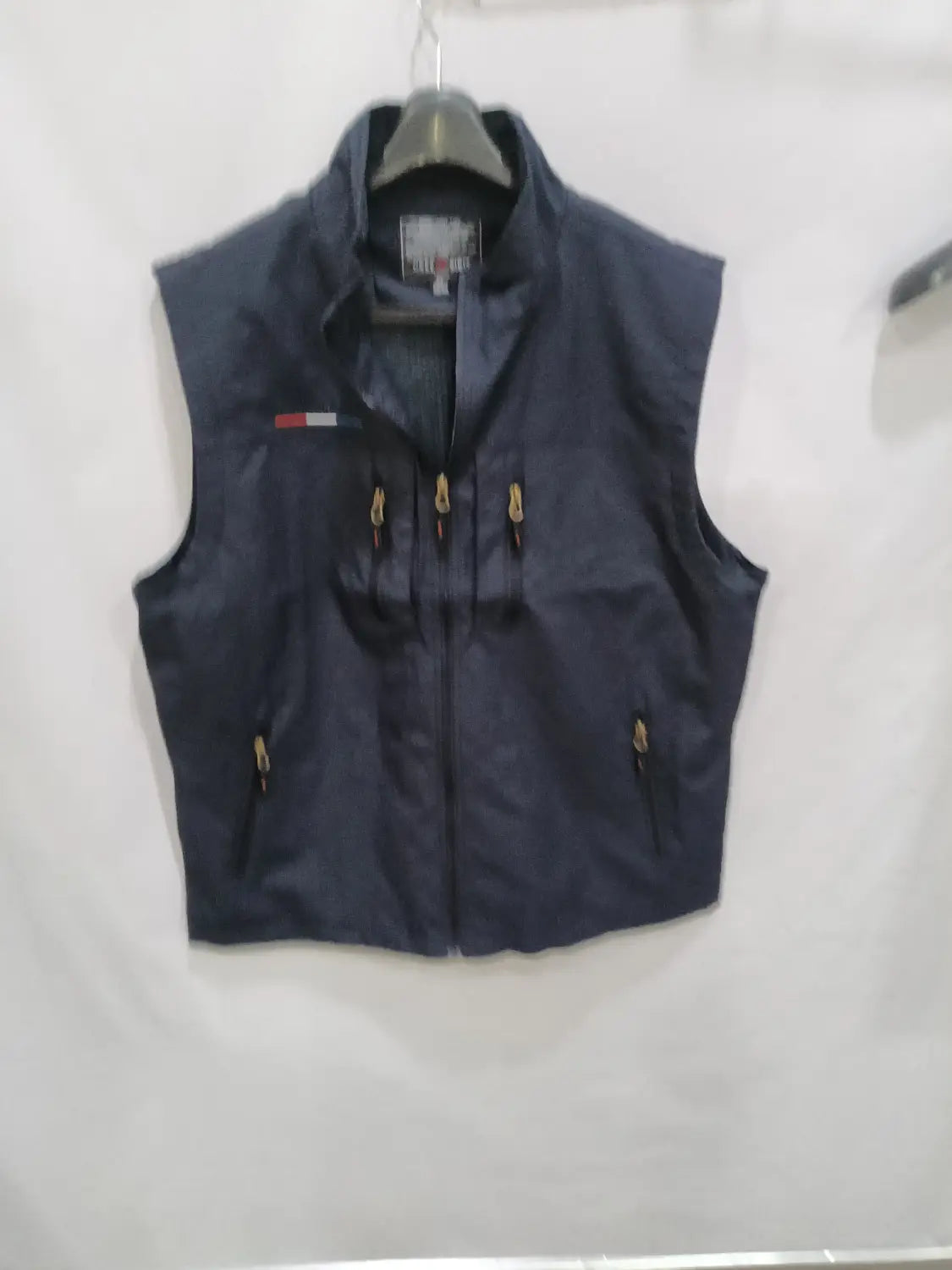 Chaleco utilitario con capucha y múltiples bolsillos para hombre en azul marino oscuro de tommy hilfiger, con cremalleras de tono dorado y un logotipo de bandera rojo, blanco y azul en el lado izquierdo del pecho.