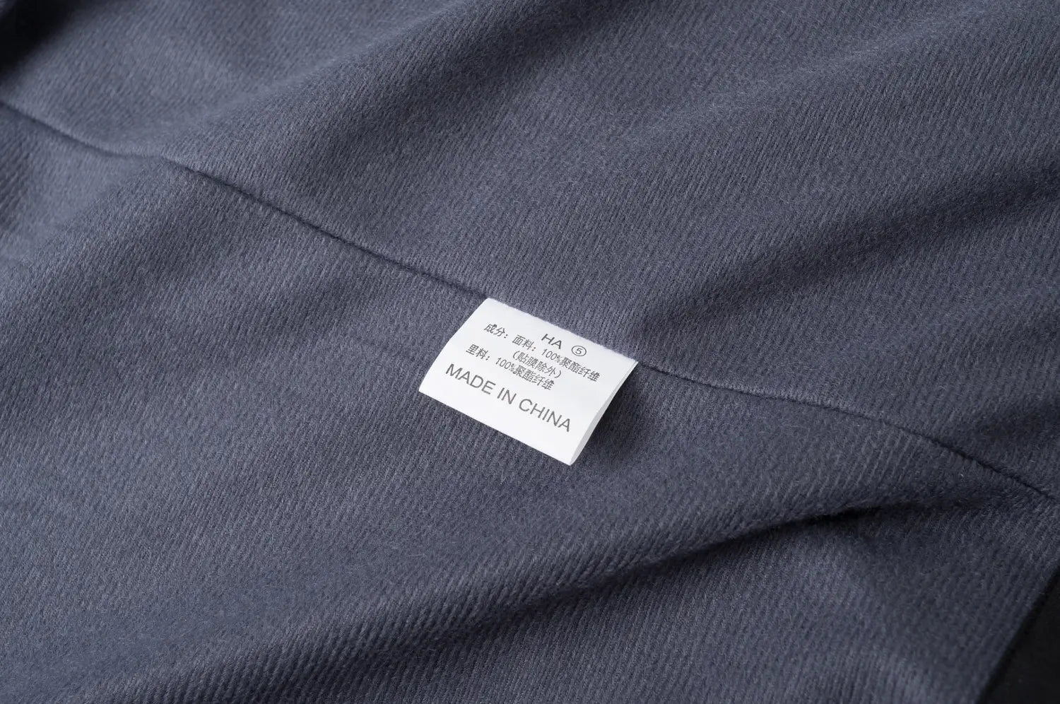 Étiquette rectangulaire d’entretien du tissu en blanc, avec le texte « Made in China » et des caractères chinois cousus sur la veste de plein air imperméable à capuche texturée gris foncé pour hommes et femmes.