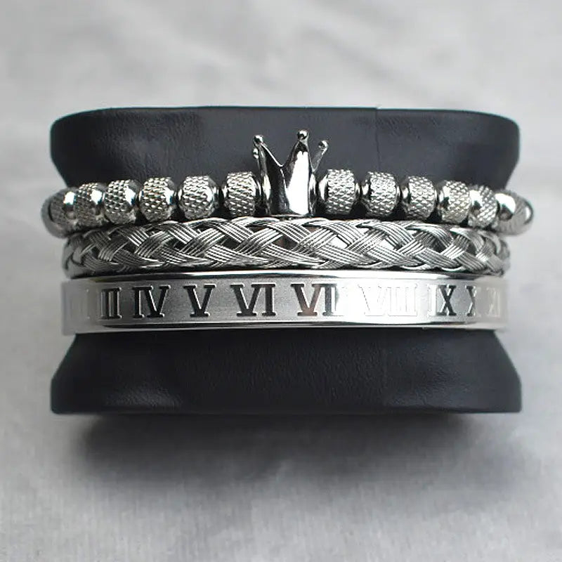 Stacked silver-toned crown royal diamond beaded bracelet set featuring braided bands textured beads with a central crown charm and an engraved roman numeral cuff.