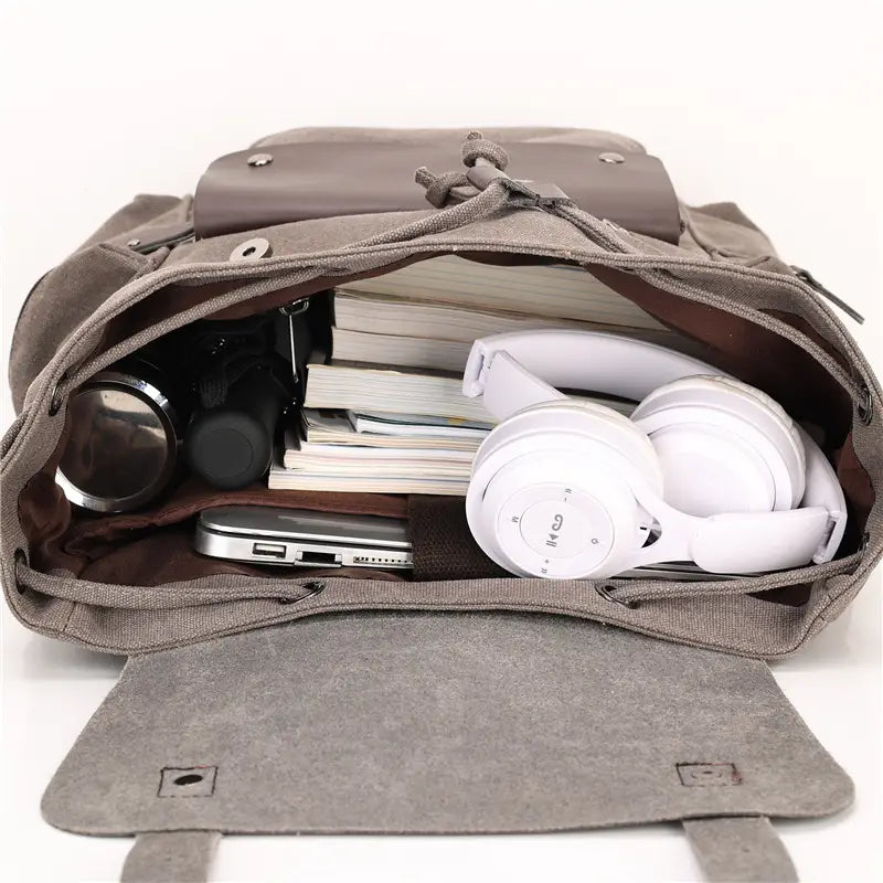 Gray canvas retro backpack with large capacity wear resistant material featuring a drawstring closure and leather-like flap filled with books headphones and tech gadgets.