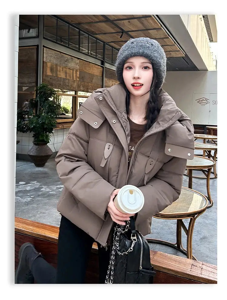 Veste courte matelassée à capuche pour femme beige avec panneaux matelassés et boutons pression à tons argentés, conçue comme un manteau d’hiver chaud.