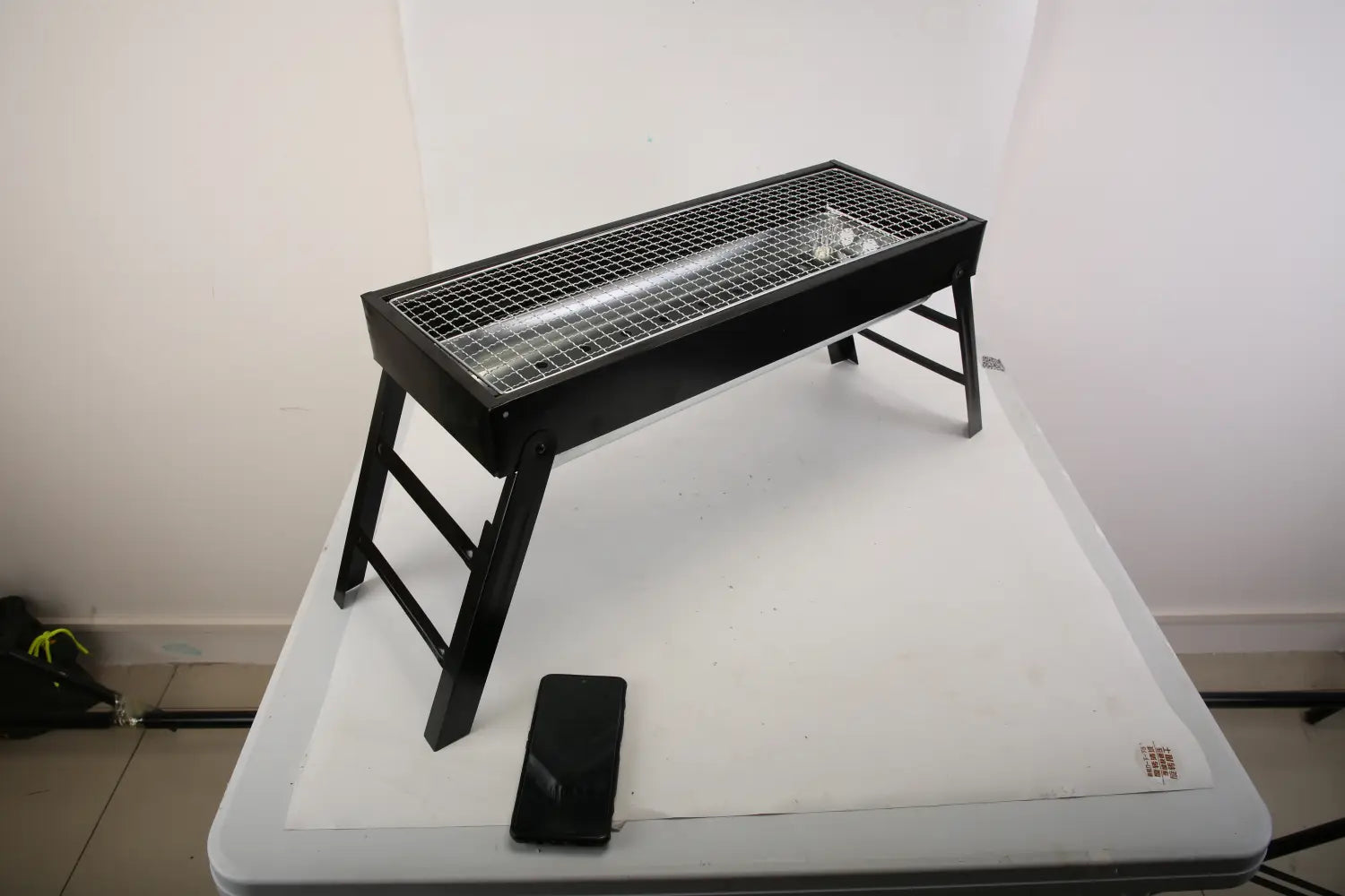 Black metal folding grill with a wire mesh cooking surface and angled legs made of stainless steel for portable charcoal barbecue use.