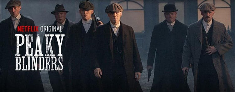 Six men dressed in dark tailored suits wearing herringbone woolen octagonal flat caps in various shades of grey and black.