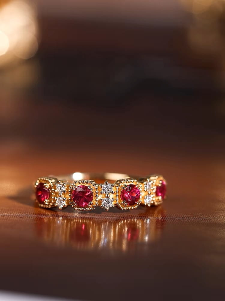 An 18k gold plated band ring featuring five round pigeon blood red rubies set in intricate metalwork with smaller clear diamonds flanking each stone.