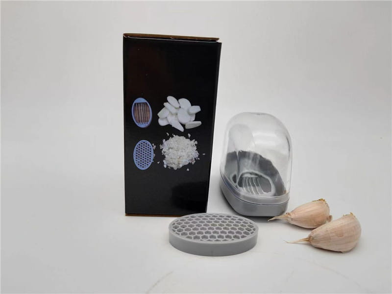 Presse-ail en plastique gris avec lames en acier inoxydable, dotée d’un couvercle en dôme transparent et d’une plaque de broyage en nid d’abeille, dans un étui gris.