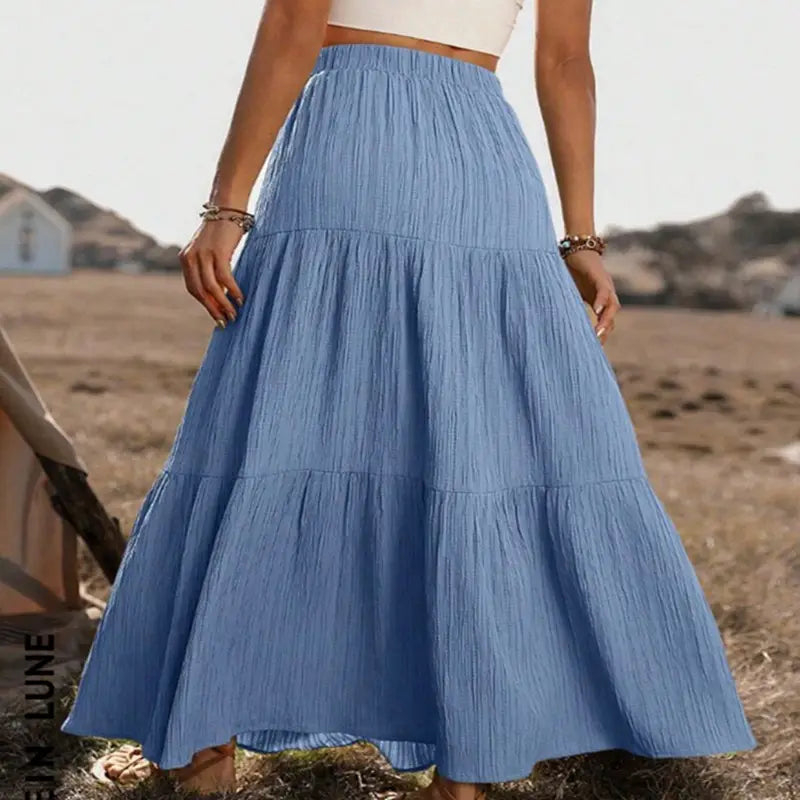 A flowing light blue tiered maxi skirt with a button front, lightweight fabric, gathered waist, and subtle vertical pleats suitable for casual wear.