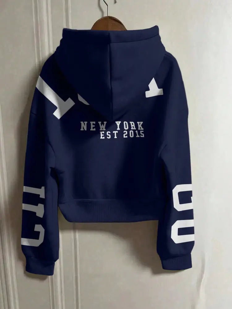 Sweat-shirt à capuche zippé court bleu marine, avec impression graphique blanche, présentant « New York Est 2015 » sur l’ensemble du dos et des numéraux audacieux sur chaque manche visibles depuis le côté arrière.