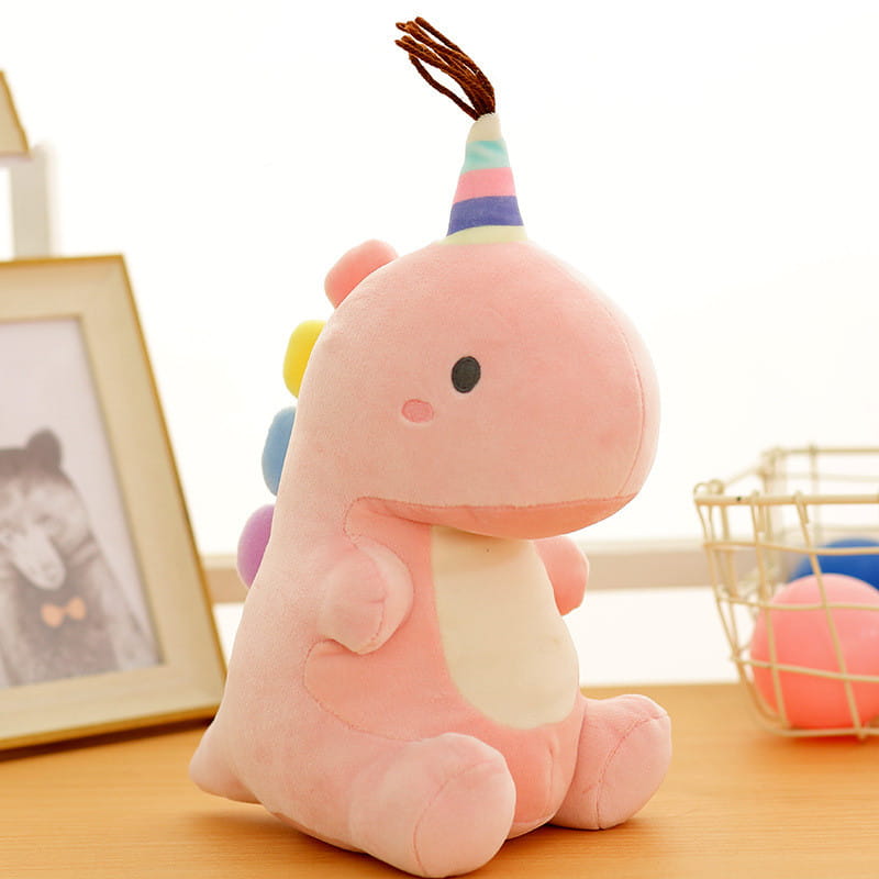 Ein pinkfarbener Dinosaurier mit einer regenbogen-gestreiften Party-Kappe mit braunen Quasten – ein weiches Kuscheltier für Spaß und Komfort.