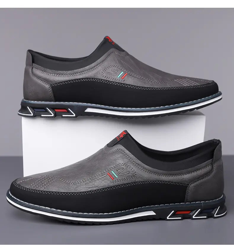 Mocasines de vestir de cuero para hombre, estilo casual de negocios, en gris y negro con parte superior texturizada, costuras contrastantes y una suela llamativa con patrón rojo y blanco.