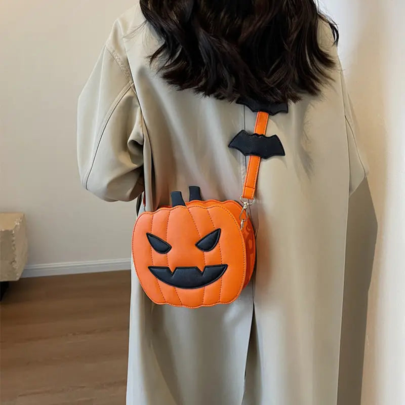 Orange pumpkin-shaped crossbody bag with black stitched facial features and a matching bat-themed strap with detachable charm.