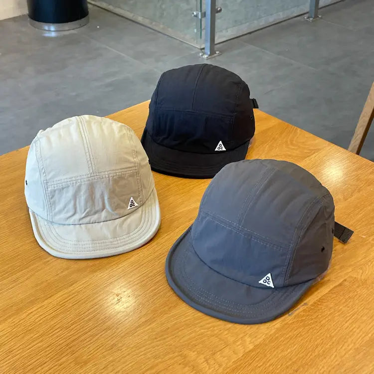 Three minimalist quick drying short brim five panel hats in cream black and gray with subtle triangular logos on the front.