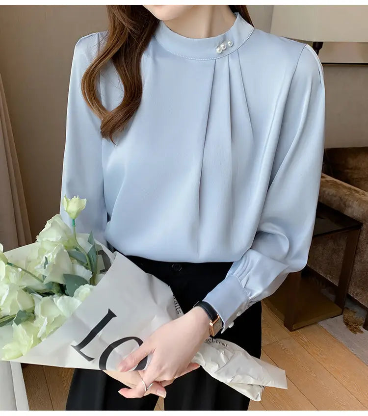 Blouse ample en satin bleu clair avec des accents de boutons perle, à encolure haute et manches longues, avec des poignets froncés.