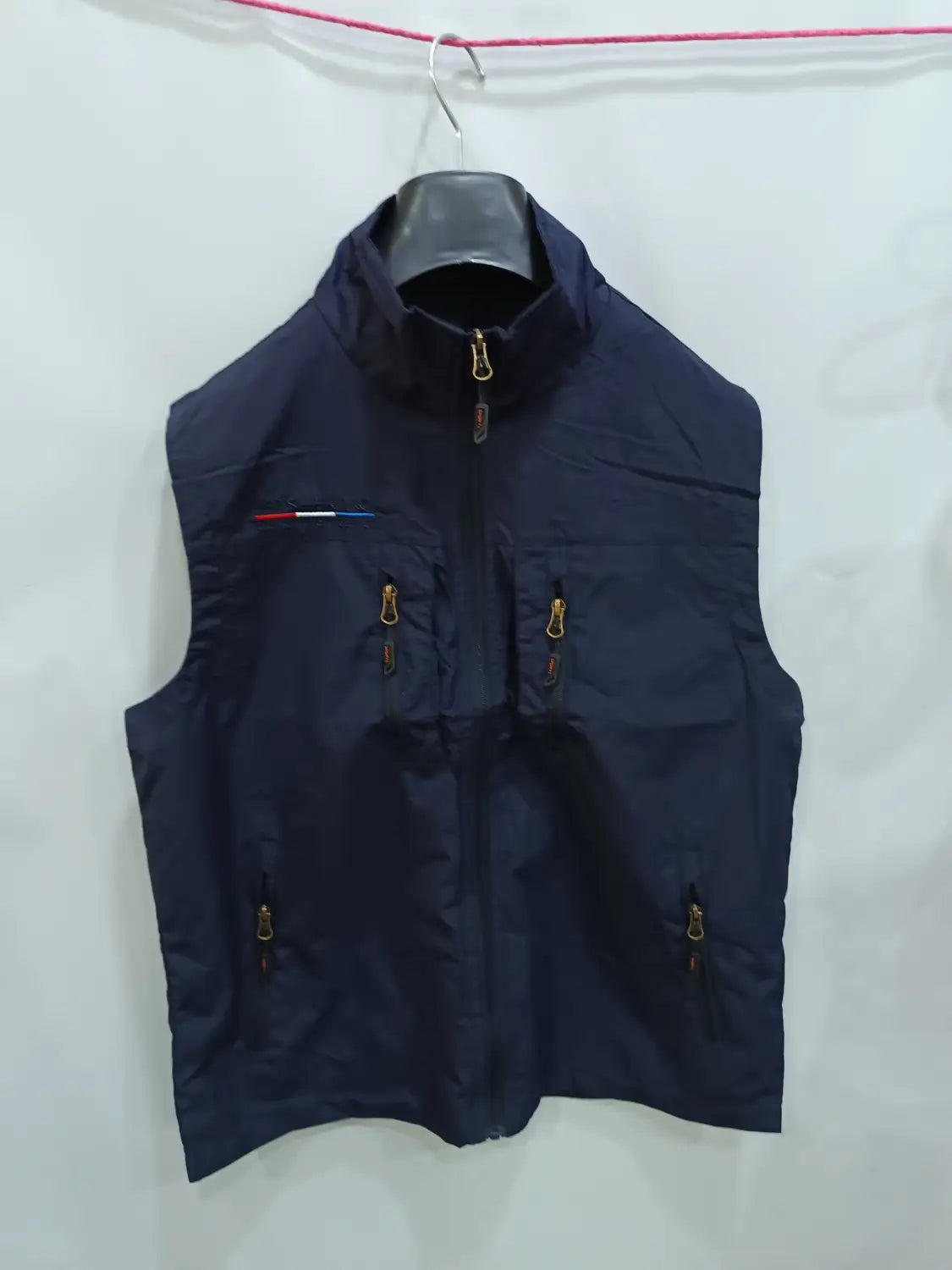 Chaleco utilitario con capucha sin mangas para hombre en azul marino oscuro, con cremalleras doradas y un detalle de franja roja, blanca y azul en el lado izquierdo del pecho.